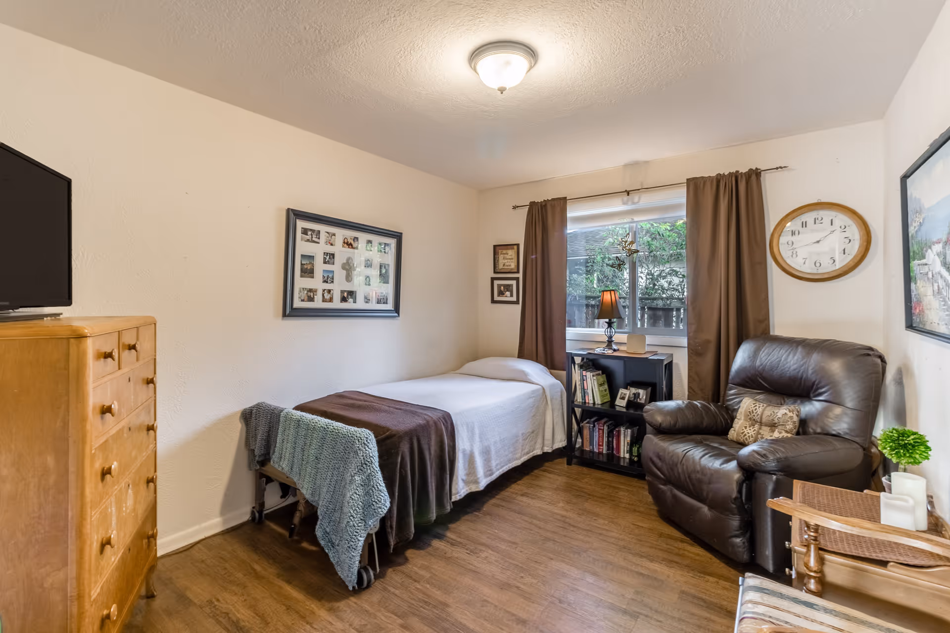 Small furnished bedroom with a single bed, leather recliner, wooden dresser, and a window with curtains.