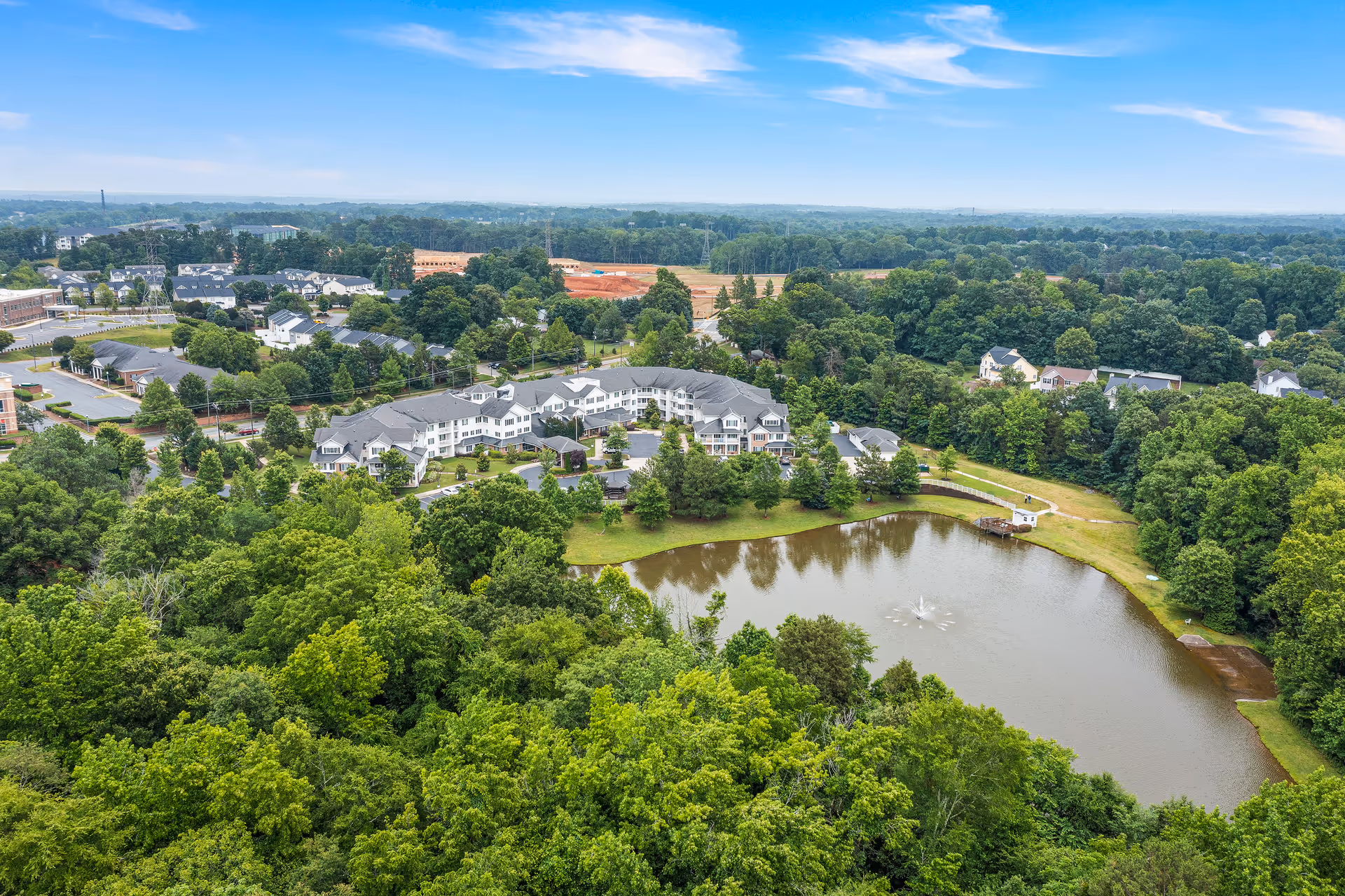 Aerial view of Shads Landing Gracious Retirement Living facility surrounded by lush green trees, with a large pond featuring a water fountain in the foreground and a clear blue sky above.