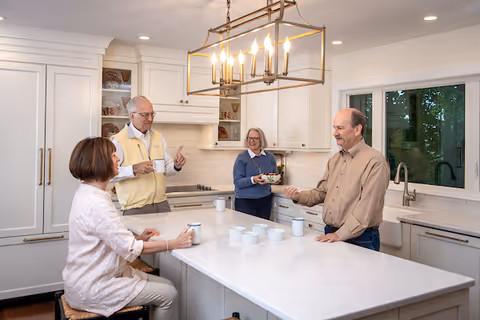 Four elderly adults socializing in a bright, modern kitchen with white cabinets and a large marble island. Three people are standing, holding cups, while one woman is seated on a stool at the island. A large rectangular chandelier with multiple lights hangs above the island, and a window shows greenery outside.