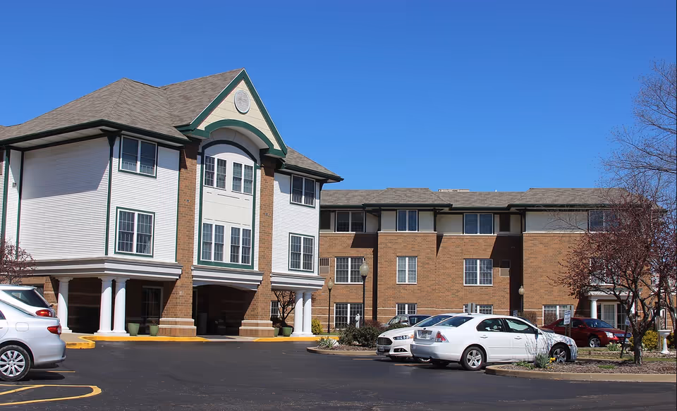 Exterior view of a senior living facility building with a combination of brick and white siding, multiple windows, and a covered entrance supported by white columns. Several cars are parked in the parking lot in front of the building under a clear blue sky.