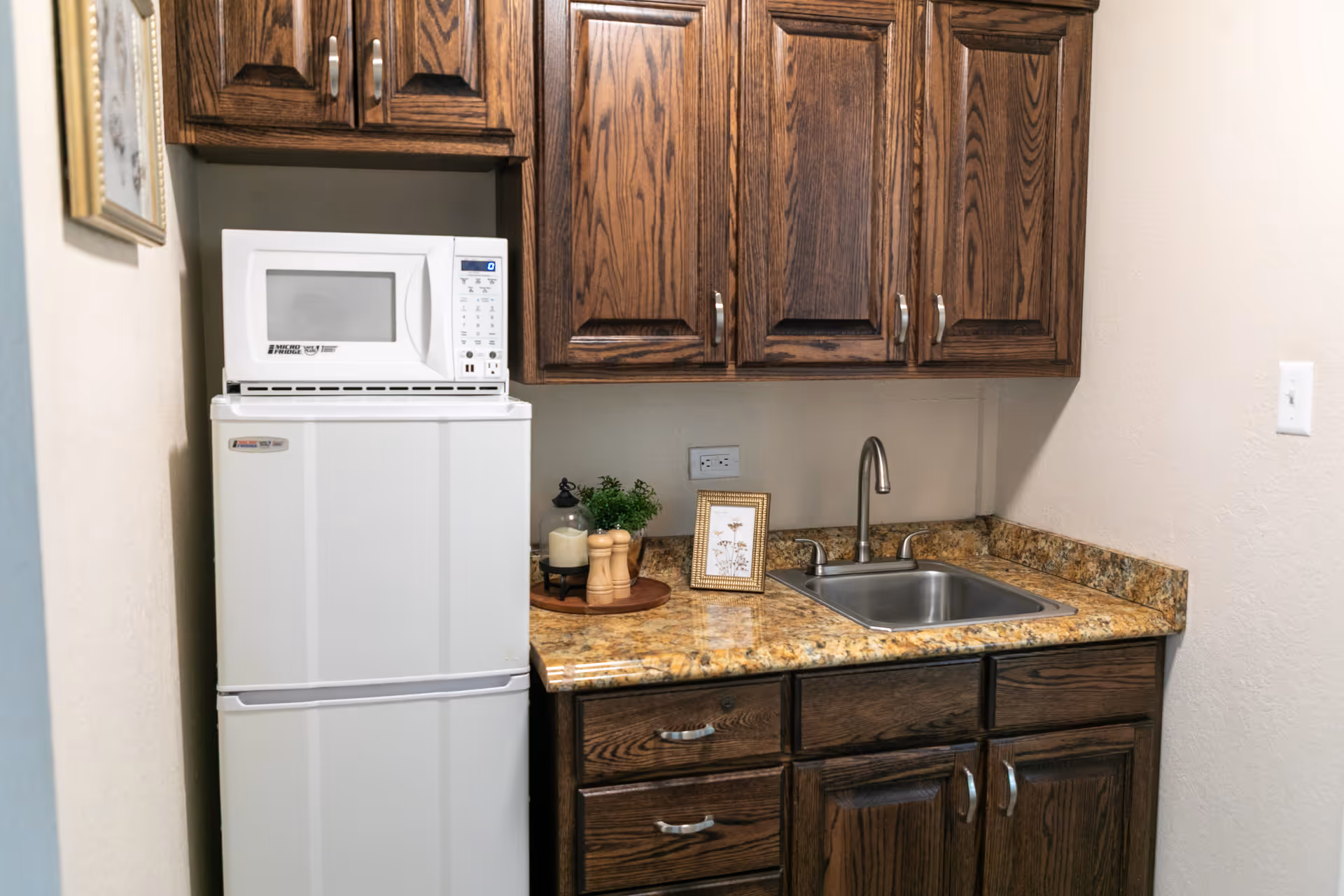 A small kitchen area with dark wooden cabinets, a granite countertop, a stainless steel sink with a faucet, a white microwave on top of a white refrigerator, and decorative items including a framed picture, a candle, and salt and pepper shakers.