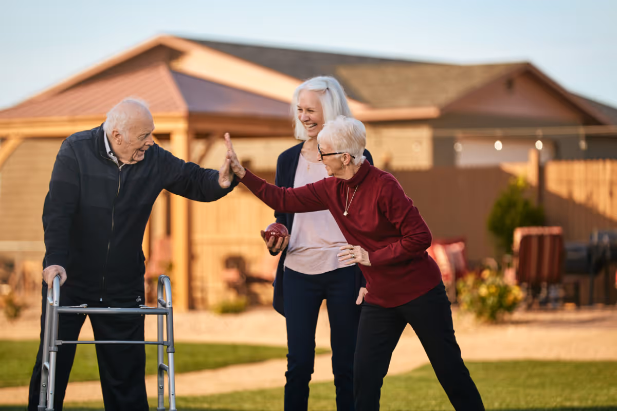 Two elderly individuals, one using a walker, giving each other a high five outdoors while a middle-aged woman stands behind them smiling. They are in a garden or yard area with a building and patio furniture in the background.