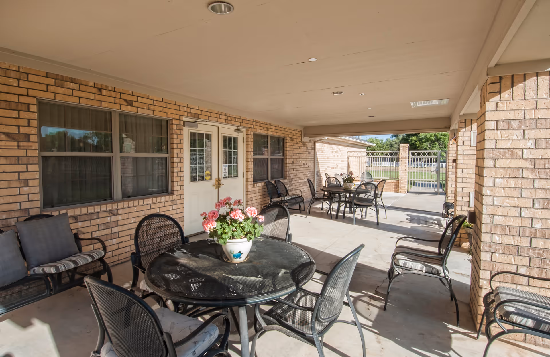 Covered outdoor patio with round tables, metal chairs, and a potted flowering plant in front of a brick building.