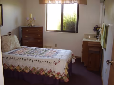 A small bedroom with a single bed covered with a patterned quilt. There is a wooden dresser with decorative candles on top against the wall near a window with a beige valance. On the right side, there is a wooden vanity with a sink and a mirror above it. A towel hangs on the wall next to the sink.