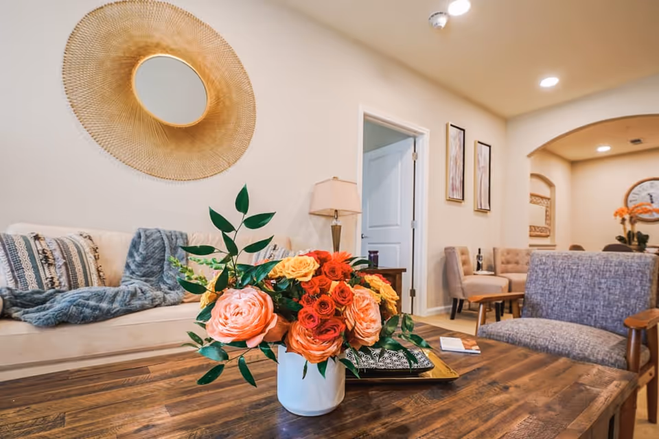 A cozy living room area with a wooden coffee table holding a vase of colorful flowers including pink, red, and yellow roses. In the background, there is a beige couch with patterned pillows and a blue throw blanket, a lamp on a side table, two framed artworks on the wall, and additional seating with upholstered chairs. The walls are light-colored and there is a decorative round mirror with a sunburst design hanging above the couch.