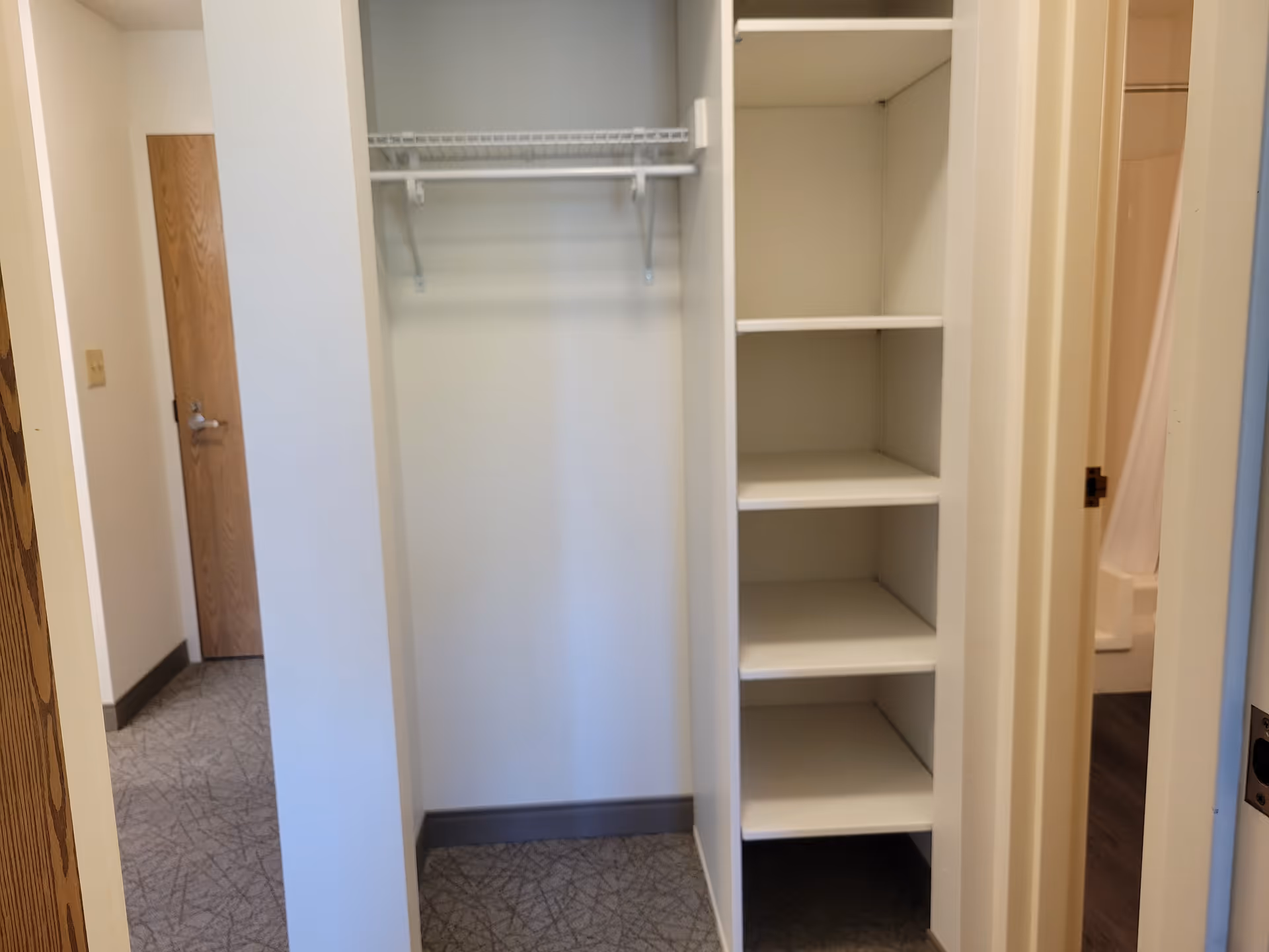 Open closet with a hanging rod and built-in shelves in an interior hallway, with a bathroom doorway visible to the right.