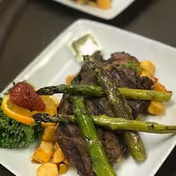 A plated meal featuring grilled asparagus spears placed on top of a serving of steak and diced potatoes, garnished with a strawberry, a slice of orange, and a piece of leafy green lettuce on a white square plate.