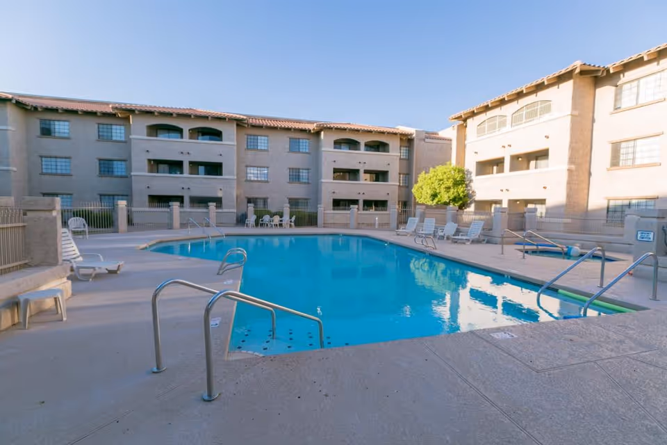Outdoor swimming pool surrounded by lounge chairs and tables, with a multi-story residential building in the background under a clear blue sky.