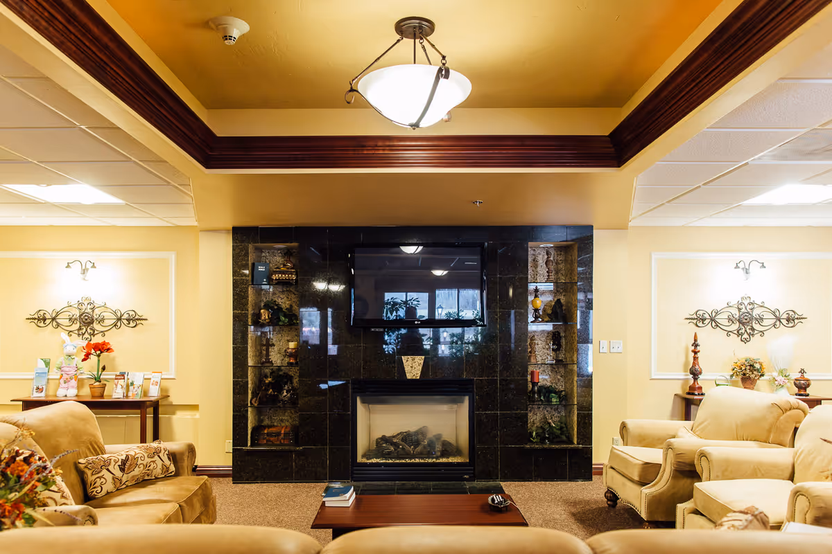 A cozy living room area with beige armchairs and sofas arranged around a wooden coffee table. The room features a black tiled fireplace with a flat-screen TV mounted above it. Decorative shelves flank the fireplace, holding various ornaments. The walls are painted a warm yellow with framed wall decor and small tables with plants and flowers on either side. A ceiling light fixture hangs above, and the ceiling has a wooden trim detail.