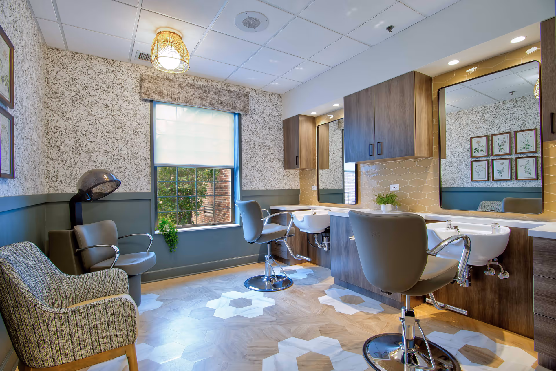 A bright and modern salon area in a senior living facility with two salon chairs in front of sinks and mirrors, a hair dryer chair, a patterned armchair, a window with a view of greenery outside, and floral wallpaper on the walls.