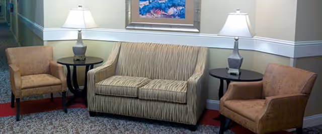 A sitting area with a beige striped loveseat flanked by two brown upholstered armchairs. Each armchair has a round black side table next to it with a table lamp on top. A framed painting hangs on the wall above the loveseat.
