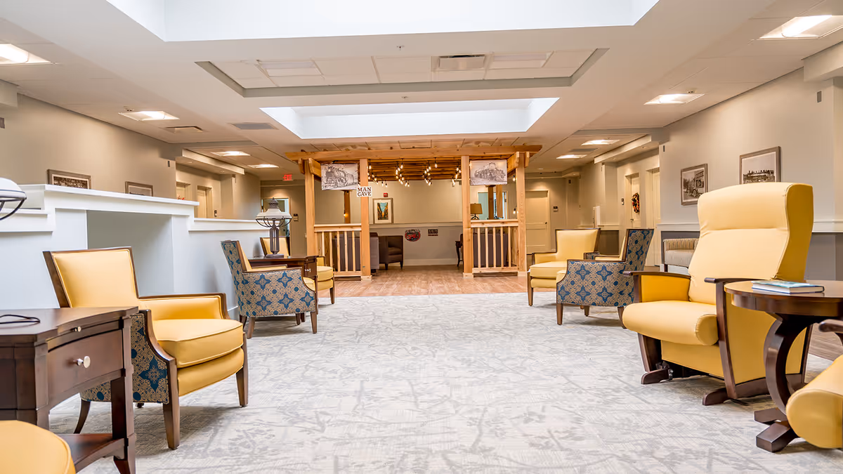 A spacious and well-lit common area in a senior living facility featuring comfortable yellow and patterned armchairs arranged around small wooden side tables. The room has a light-colored carpet and neutral walls adorned with framed pictures. At the far end, there is a wooden structure labeled 'Man Cave' with additional seating inside.