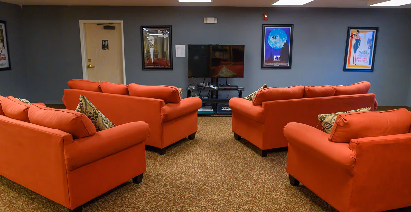 A cozy lounge area with four red sofas arranged around a flat-screen TV mounted on a black stand. The room has a carpeted floor and gray walls adorned with framed movie posters including The Godfather, E.T., and Breakfast at Tiffany's. A door is visible on the left side of the wall.