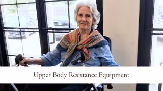 An elderly woman using upper body resistance exercise equipment in a well-lit room with large windows.