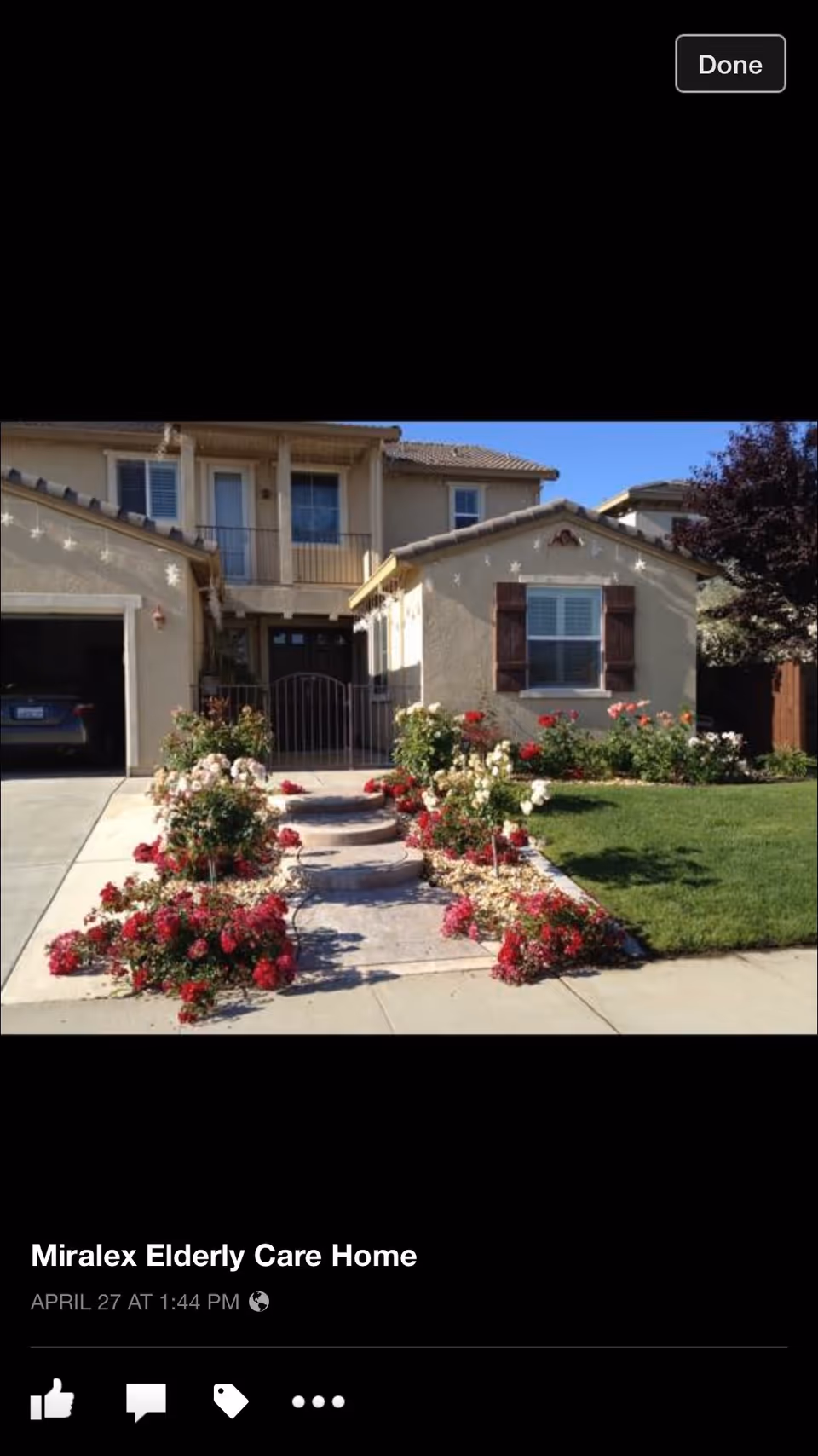 Front exterior view of a two-story beige house with a tiled roof, a small balcony, and a garden pathway lined with red and white flowers leading to the front door. There is a green lawn on the right side and a garage on the left side.