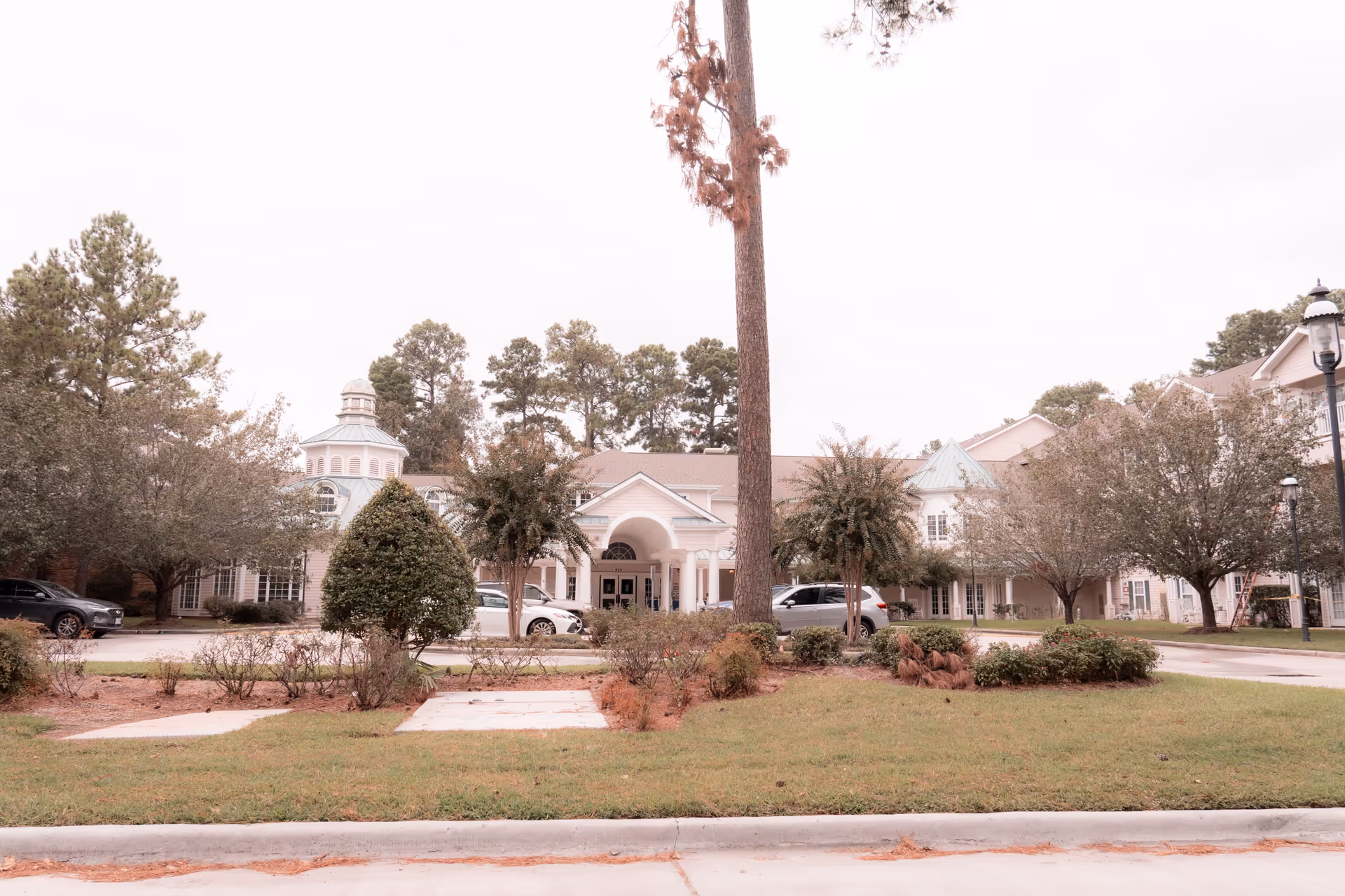 Front exterior view of King's Preserve at Kingwood senior living facility with a driveway, several parked cars, landscaped bushes, trees, and a building with a covered entrance and cupola.