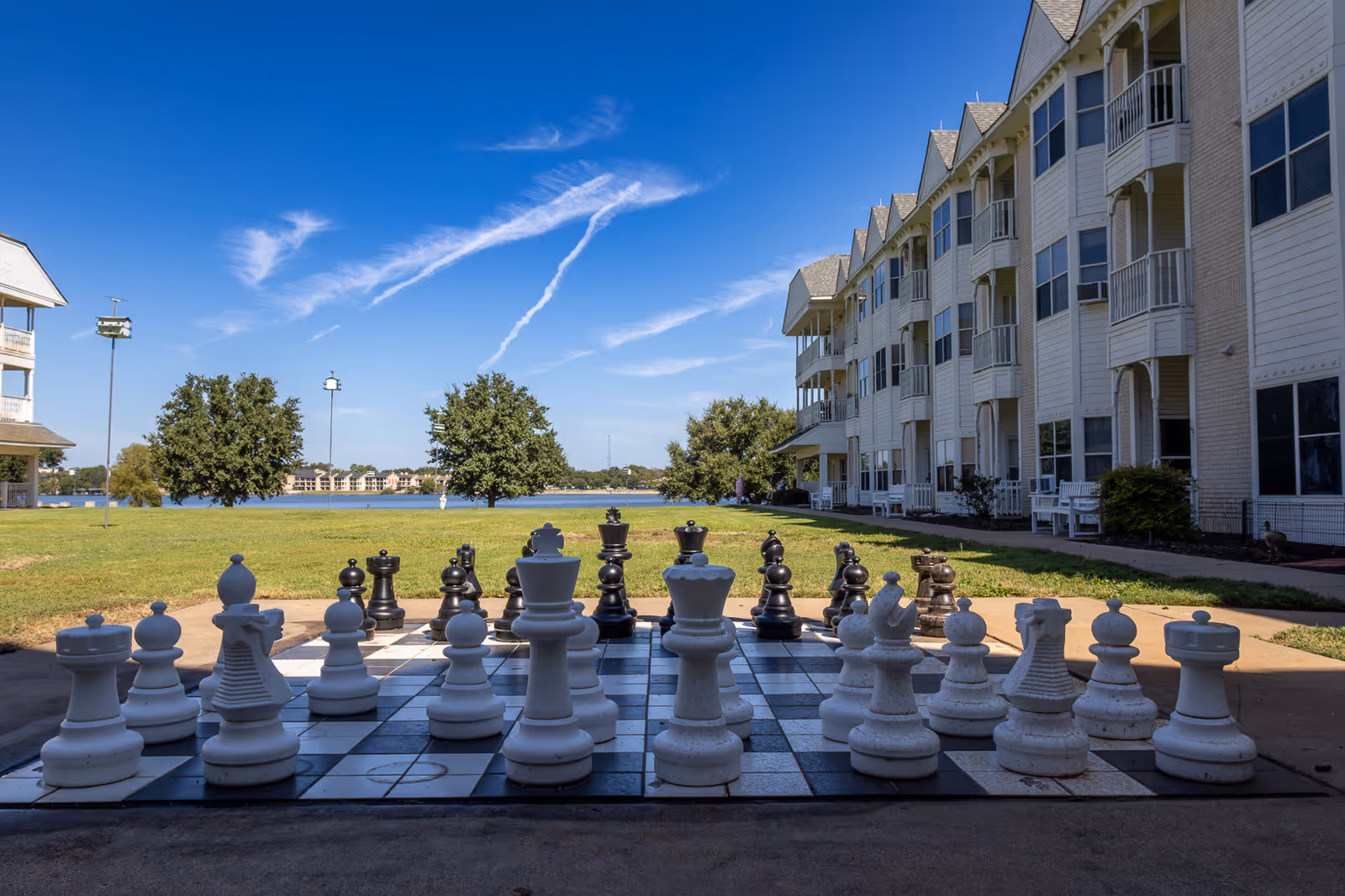 Large outdoor chessboard with oversized chess pieces set up on a paved area in front of a multi-story senior living building. Green grass, trees, and a body of water are visible in the background under a clear blue sky.