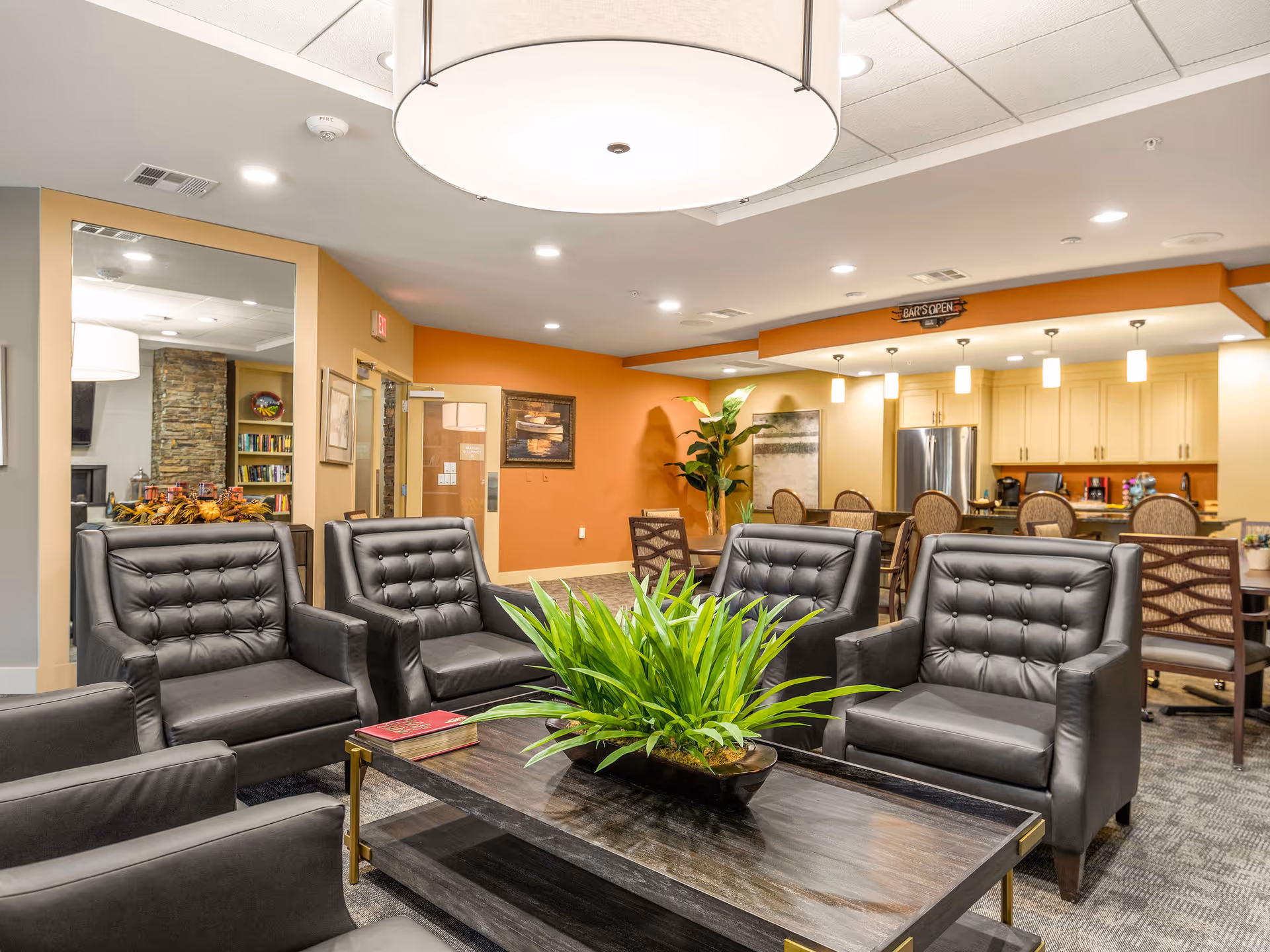 Bright communal lounge with leather armchairs arranged around a coffee table holding a green plant, with a kitchenette and dining area in the background.