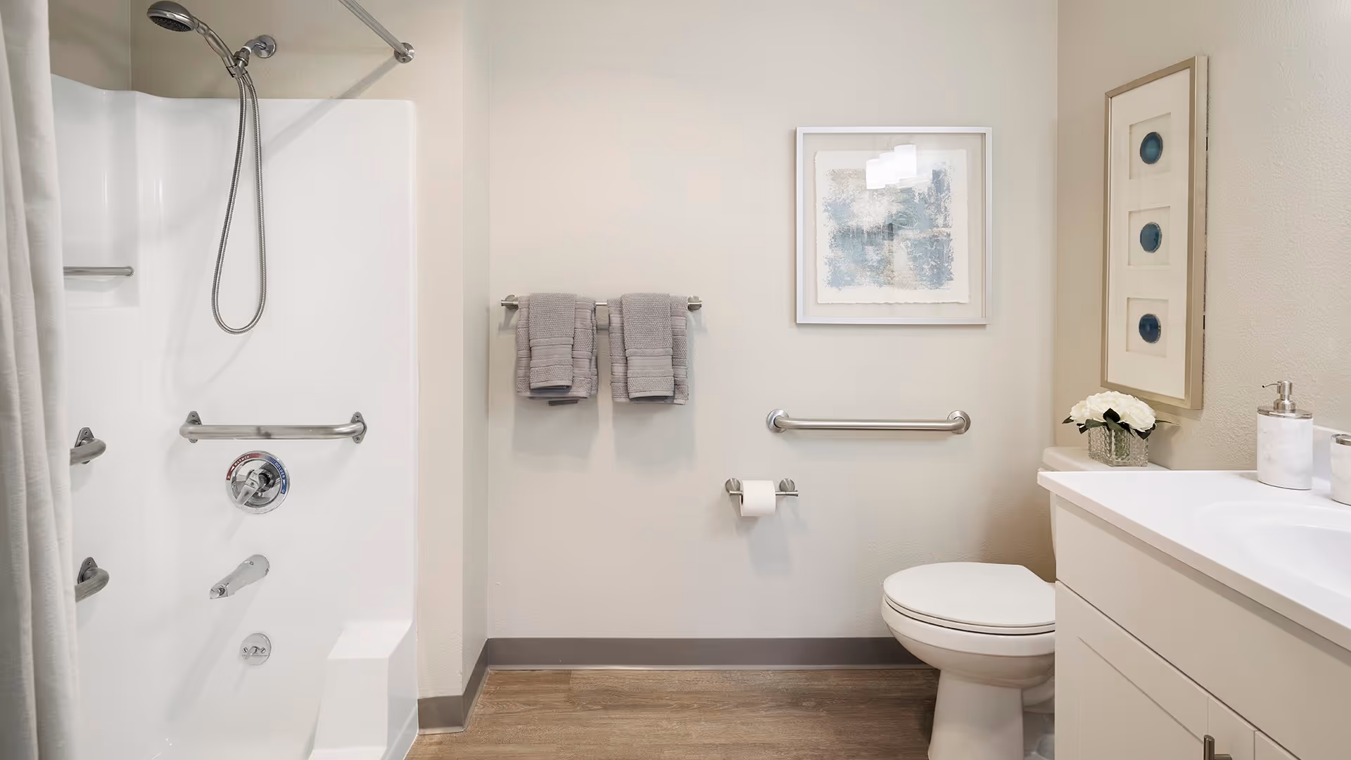 A clean and modern bathroom featuring a white bathtub with a handheld showerhead and grab bars, a toilet with a grab bar beside it, a white vanity with a sink, soap dispenser, and a small vase of white flowers. Two gray towels hang on a towel rack, and two framed artworks are displayed on the walls. The floor has a wood-like finish.