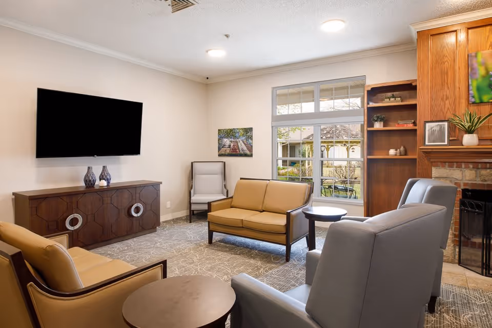 A cozy living room in a memory and dementia care facility featuring a wall-mounted flat-screen TV above a wooden cabinet with decorative vases. The room has a beige loveseat, two gray armchairs, and a beige armchair arranged around small round tables. There is a large window showing an outdoor gazebo and garden area. A wooden bookshelf and a brick fireplace with a wooden mantel are also visible.