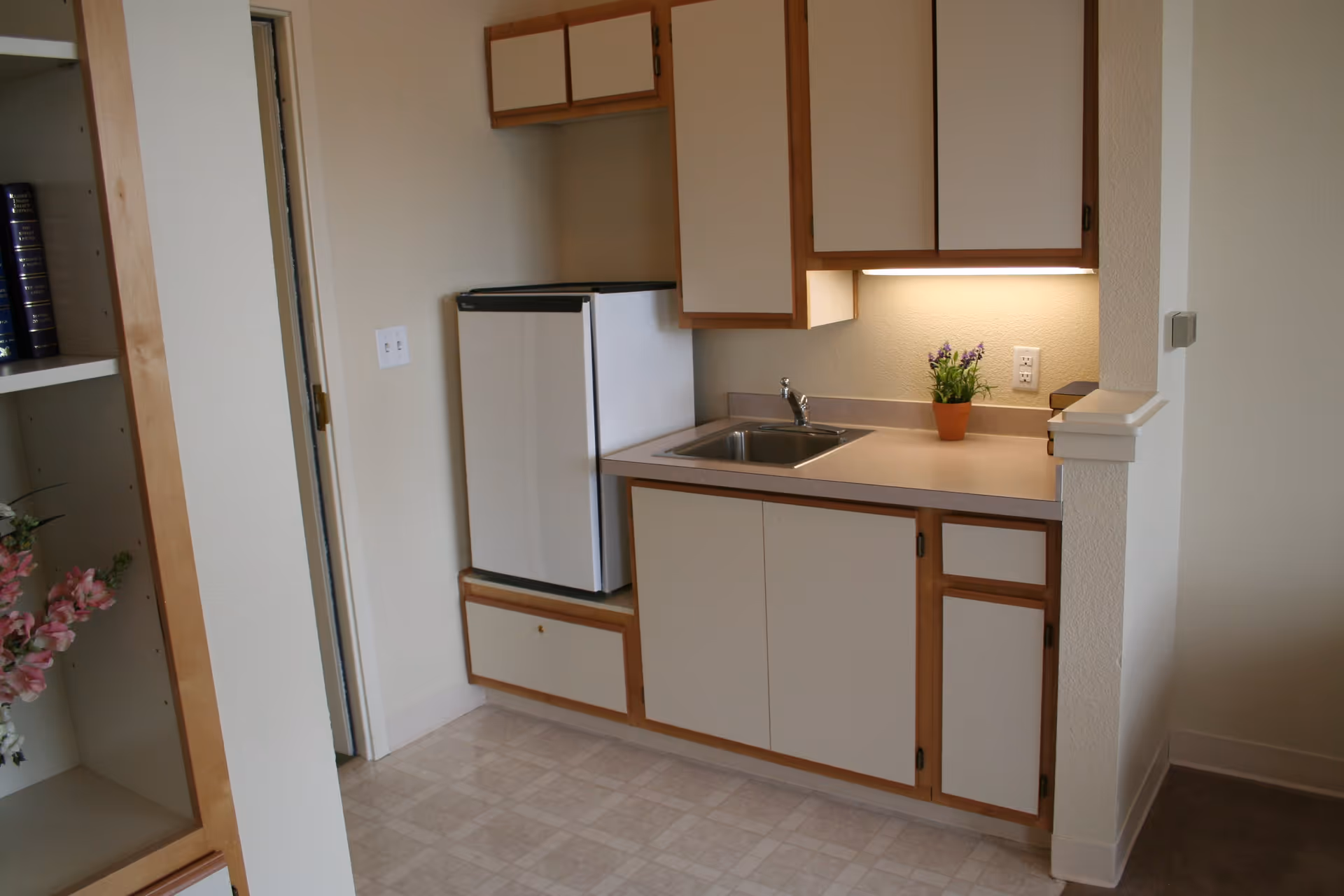 Small kitchenette area with a compact refrigerator, a sink, upper and lower cabinets, and a small potted plant on the countertop. The kitchenette is adjacent to a built-in shelving unit with books and flowers.