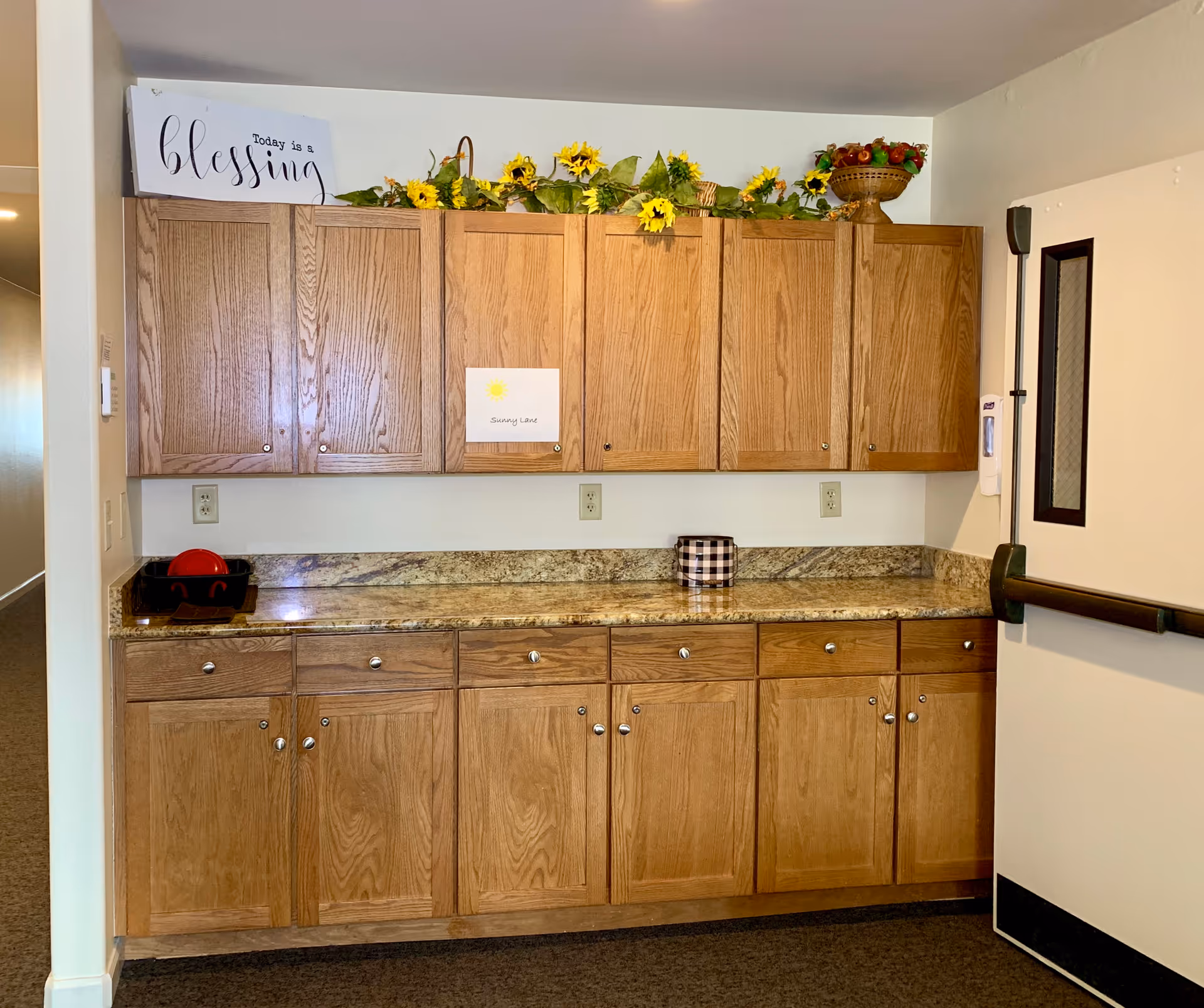 A kitchen area with wooden cabinets above and below a granite countertop. On top of the upper cabinets, there is a decorative arrangement of sunflowers and a bowl of artificial fruit. A sign on the wall reads 'Today is a blessing' and another smaller sign says 'Sunny Lane' with a drawing of a sun. To the right, there is a door with a window and a hand sanitizer dispenser mounted on the wall.
