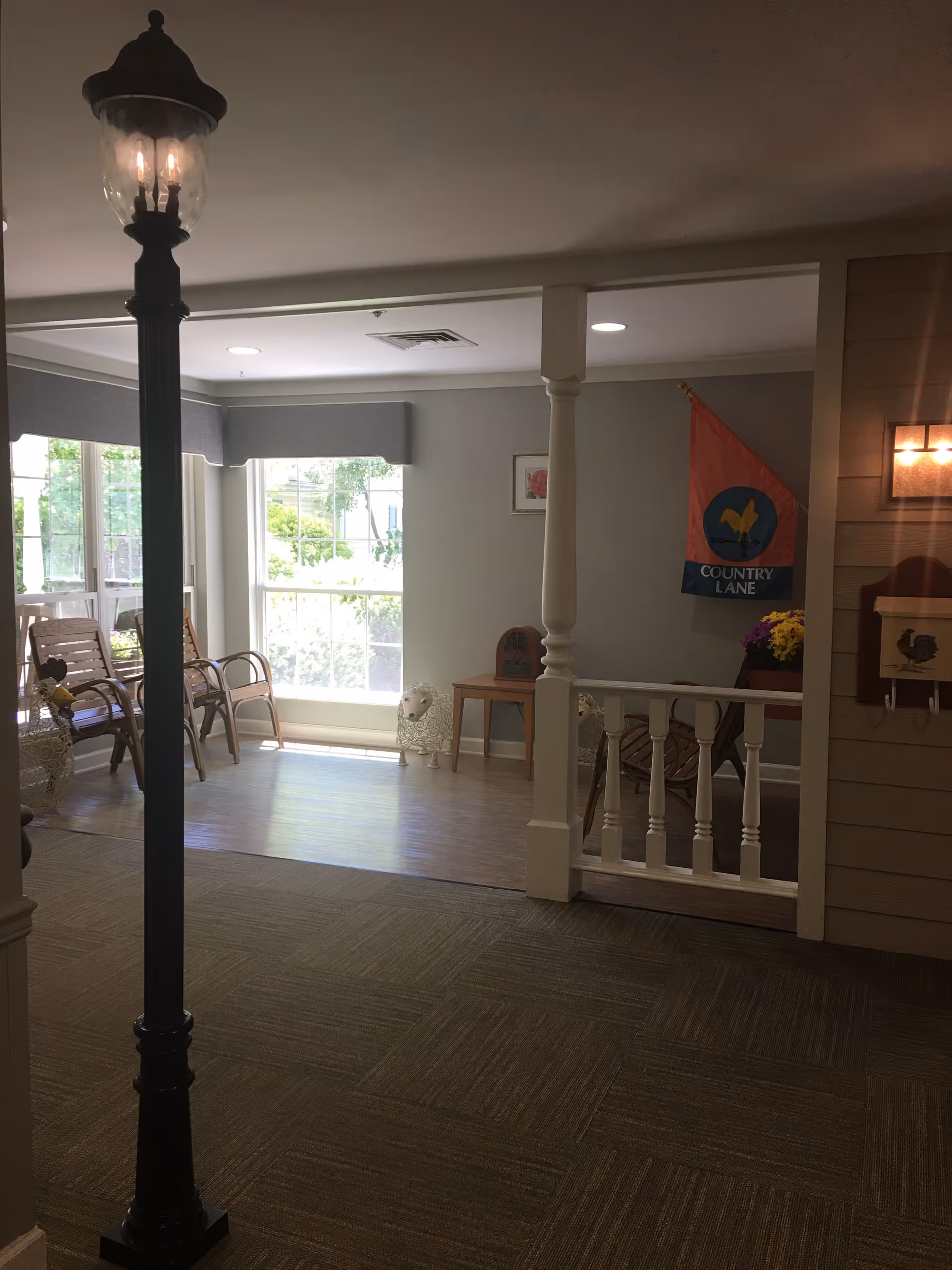 Interior common area with a decorative street lamp, chairs by a large window, small tables and a 'Country Lane' banner on the wall.