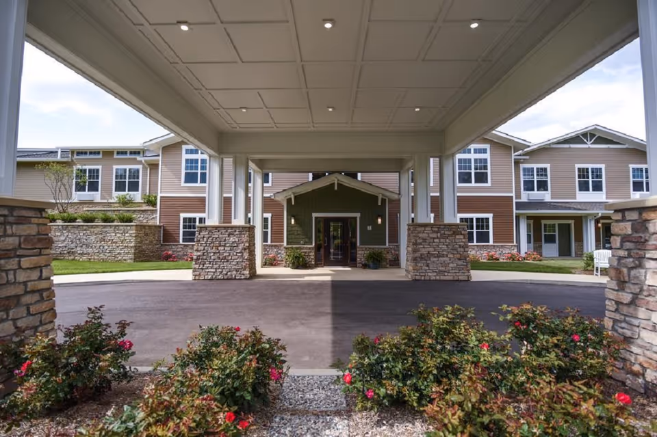 View of the entrance to a senior living facility named Traditions of Beavercreek, showing a covered driveway with stone pillars, landscaped flower beds with red flowers, and a multi-story building with beige and brown siding and multiple windows in the background.