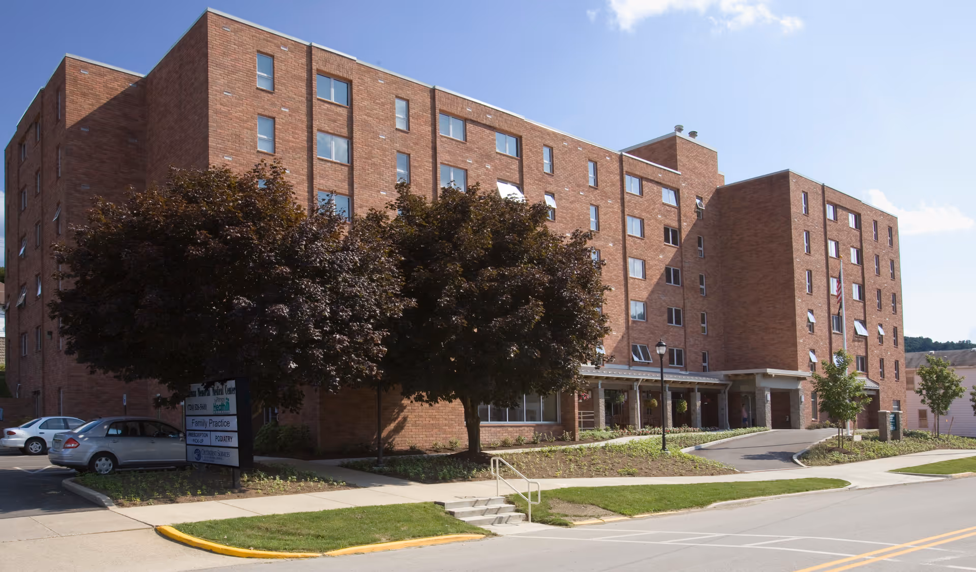 Exterior view of a multi-story brick building with several windows, a covered entrance, and a few trees and parked cars in front under a clear blue sky.