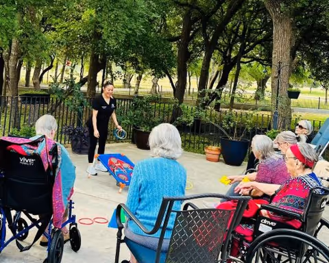 A caregiver leads a group of elderly residents in wheelchairs playing a cornhole-style game on an outdoor patio surrounded by trees.
