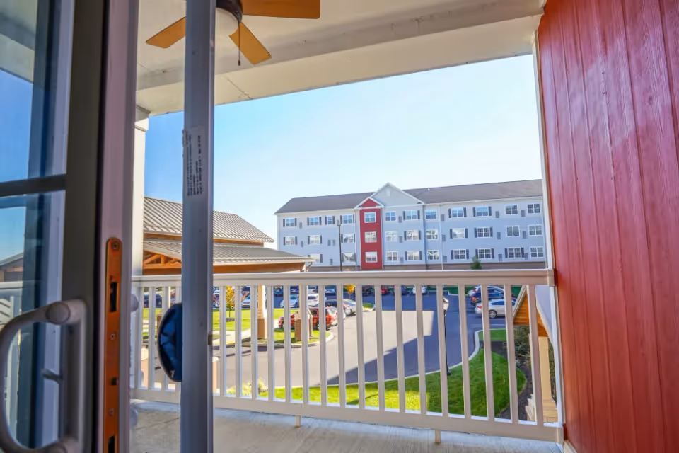 View from a balcony through a sliding glass door overlooking a courtyard and the front of a multi-story senior living building with white siding and a red central accent.