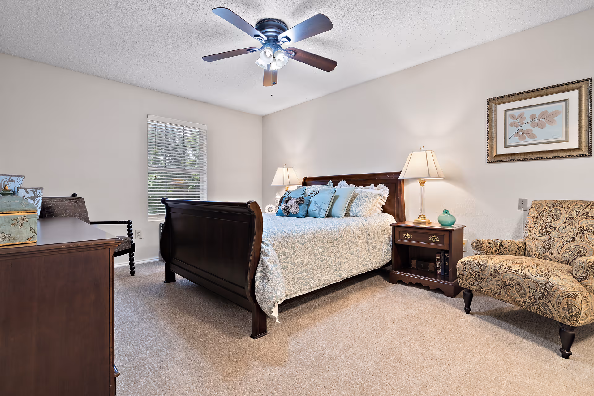 A cozy bedroom with a dark wooden bed frame, a bed with light-colored patterned bedding and multiple pillows, a nightstand with a lamp and decorative items, a patterned armchair, a wooden dresser, a window with blinds, and a ceiling fan with lights.