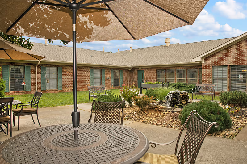 Outdoor courtyard area at The Heritage Center featuring a round metal table with an umbrella, several metal chairs, a small rock garden with a water fountain, and a brick building with green window shutters in the background under a partly cloudy sky.