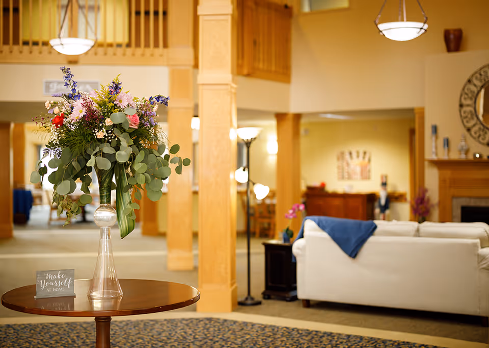 A bright and welcoming senior living facility common area with a large floral arrangement on a round wooden table in the foreground. The background features a white couch with a blue throw, wooden pillars, a floor lamp, and warm yellow walls with decorative elements including a mirror and artwork.