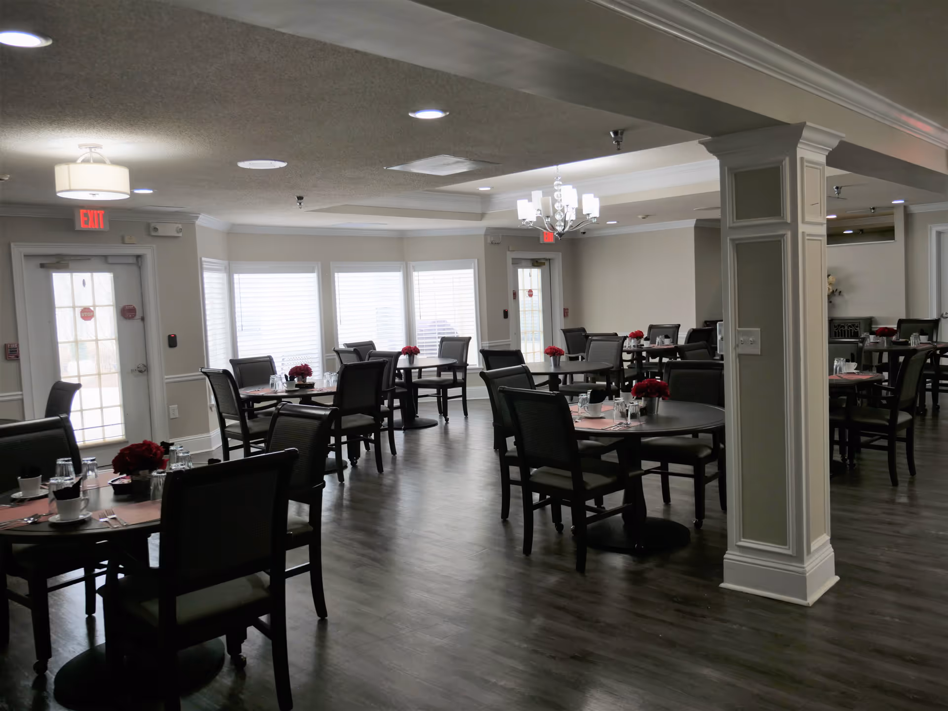 A dining room with multiple round tables, each set with placemats, cups, glasses, and red flower centerpieces. The room has dark wood flooring, beige walls, white trim, and large windows letting in natural light. There are exit signs above two doors and a chandelier hanging from the ceiling.