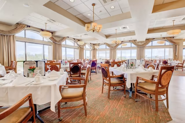 Bright and spacious dining room with large windows letting in natural light. Tables are set with white tablecloths, napkins, cups, and menus. Wooden chairs with cushioned seats surround the tables. The ceiling features recessed lighting and chandeliers, and the room has beige curtains and carpeted flooring.