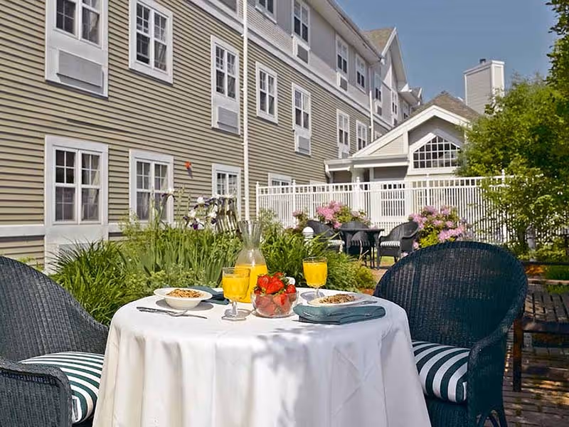 Outdoor patio area at Atria Kennebunk with a round table covered with a white tablecloth, set with two glasses of orange juice, a bowl of strawberries, and two bowls of cereal. The table is surrounded by wicker chairs with striped cushions. In the background, there is a garden with green plants and flowers, a white fence, and the exterior of a multi-story building with multiple windows.