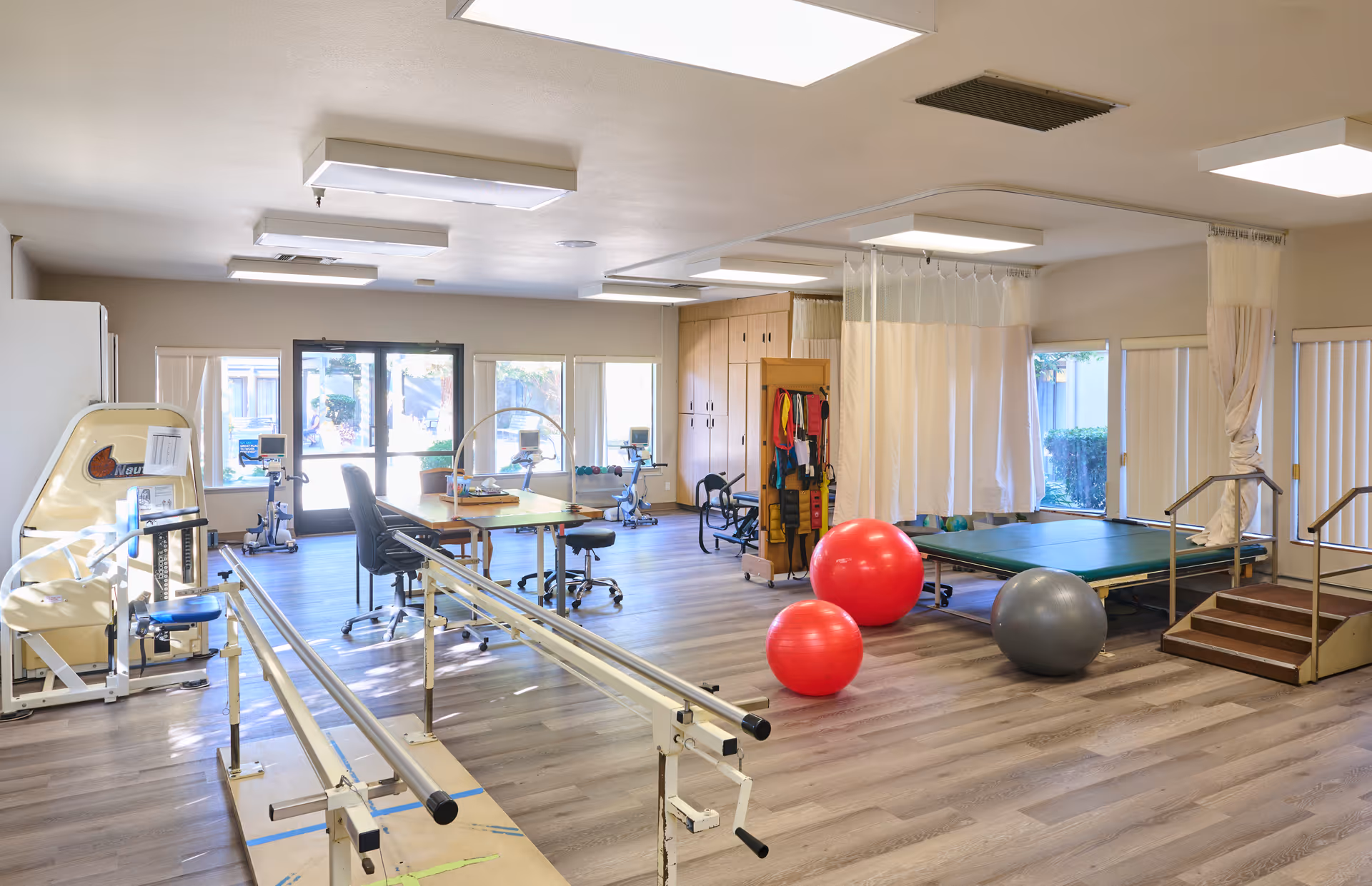 A spacious rehabilitation room with parallel bars for walking exercises, exercise balls, therapy tables with privacy curtains, exercise equipment, chairs, and large windows letting in natural light.