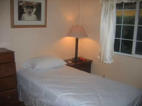 Small bedroom with a single bed, nightstand lamp, dresser, and window curtained.
