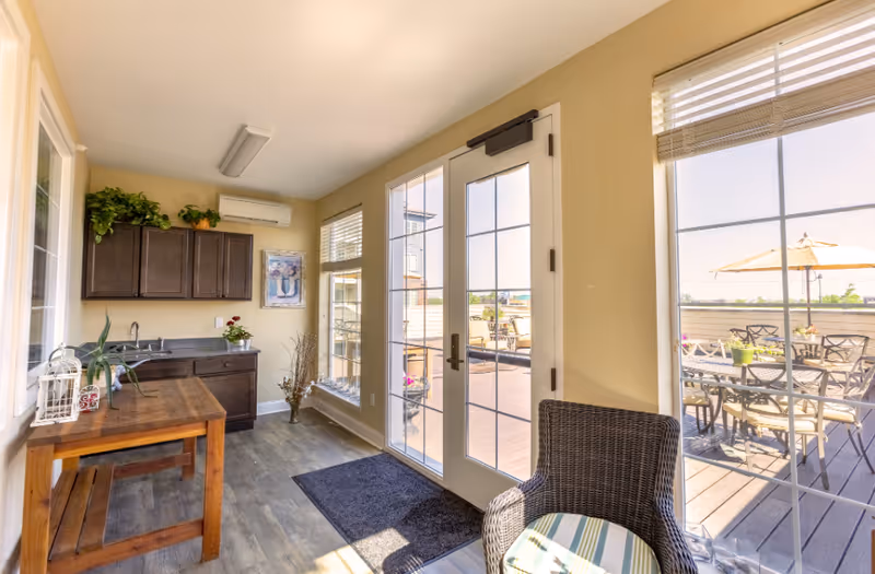 A bright interior room with wooden flooring, featuring a small kitchenette with dark cabinets, a wooden table, and a wicker chair with a striped cushion. Large glass doors and windows provide a view of an outdoor patio area with tables, chairs, and umbrellas.