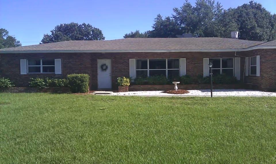 Single-story brick building with a gray shingled roof, white door with a wreath, and multiple windows with white shutters. The front yard has green grass, bushes, a small birdbath, and a lamp post. Trees are visible in the background under a clear blue sky.