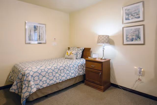 A small bedroom with a single bed covered in a blue and white patterned bedspread. Next to the bed is a wooden nightstand with a lamp, a clock, and some small items. The walls are light-colored and decorated with three framed pictures.