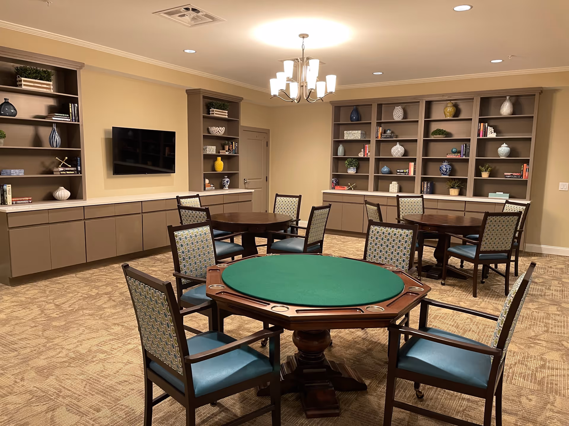 A well-lit common room with three round tables surrounded by chairs. One table in the foreground has a green felt top, likely for card games. The walls have built-in shelves decorated with books, plants, and vases. A flat-screen TV is mounted on one wall, and a modern chandelier hangs from the ceiling.
