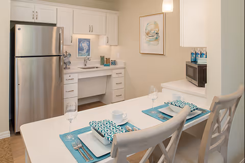 A small kitchen area with a stainless steel refrigerator, white cabinets, a sink, and a microwave on the counter. In the foreground, there is a dining counter set with two place settings including plates, bowls, utensils, cups, and wine glasses on turquoise placemats. Two wooden chairs with cross-back design are positioned at the counter. The walls are beige with framed artwork and a few bottles of water on the counter near the microwave.