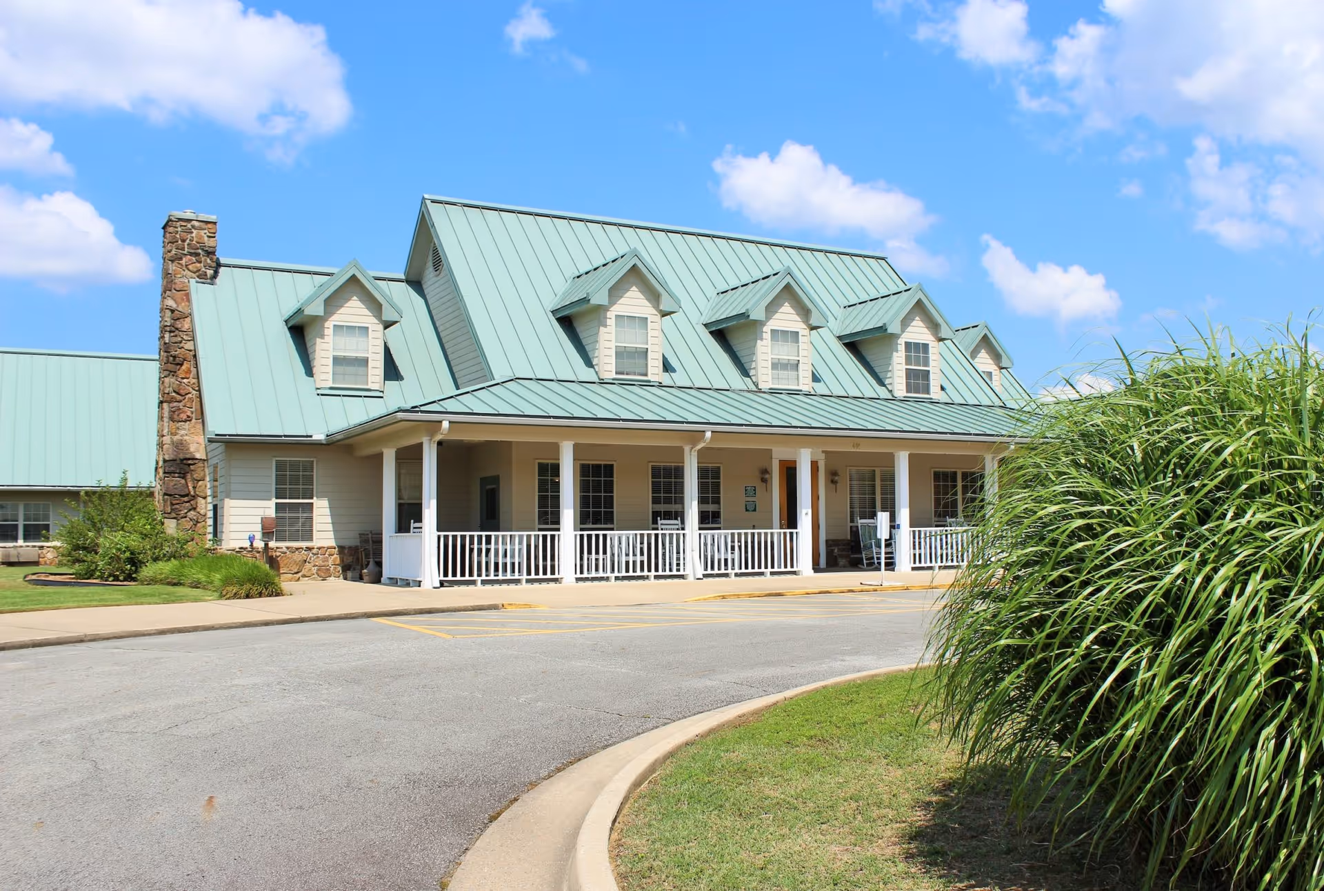 Front exterior view of a senior living facility building with a light green metal roof, multiple dormer windows, a stone chimney, and a covered porch with white railings. The building is surrounded by a paved driveway and green landscaping under a blue sky with scattered clouds.