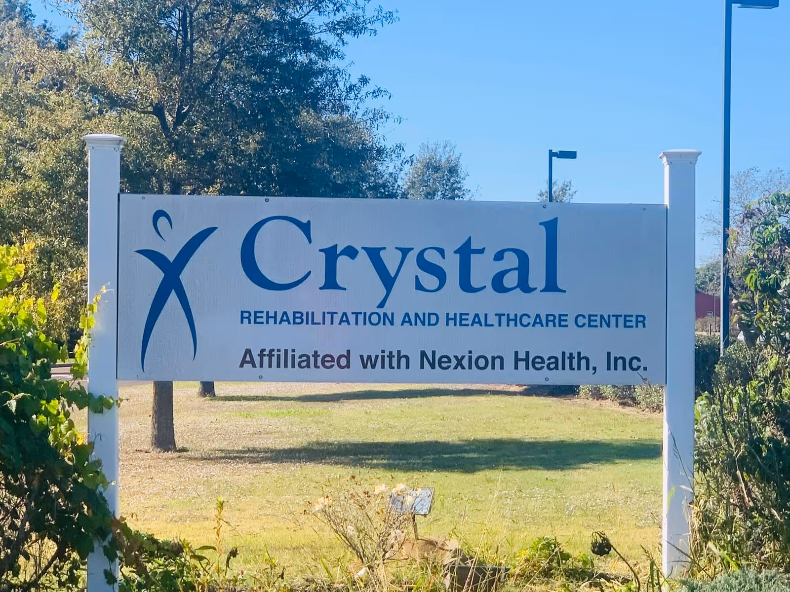 Outdoor sign for Crystal Rehabilitation and Healthcare Center, affiliated with Nexion Health, Inc., standing on a grassy area with trees and bushes in the background under a clear blue sky.