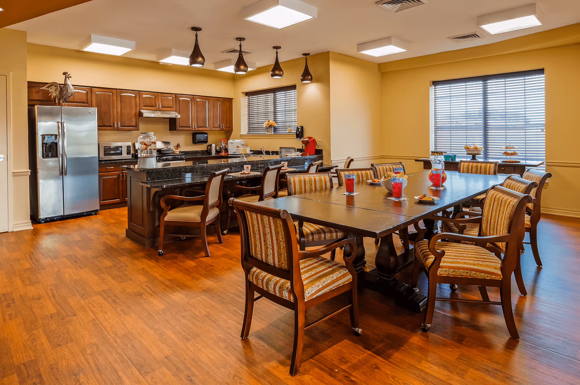 A spacious kitchen and dining area with wooden floors and yellow walls. The kitchen features dark wood cabinets, a stainless steel refrigerator, a microwave, and a stove with a range hood. A large island with a dark granite countertop has several chairs around it. The dining area has a large wooden table surrounded by cushioned chairs with striped upholstery. The table is set with glasses of red drink and plates with snacks. Two windows with blinds allow natural light into the room.