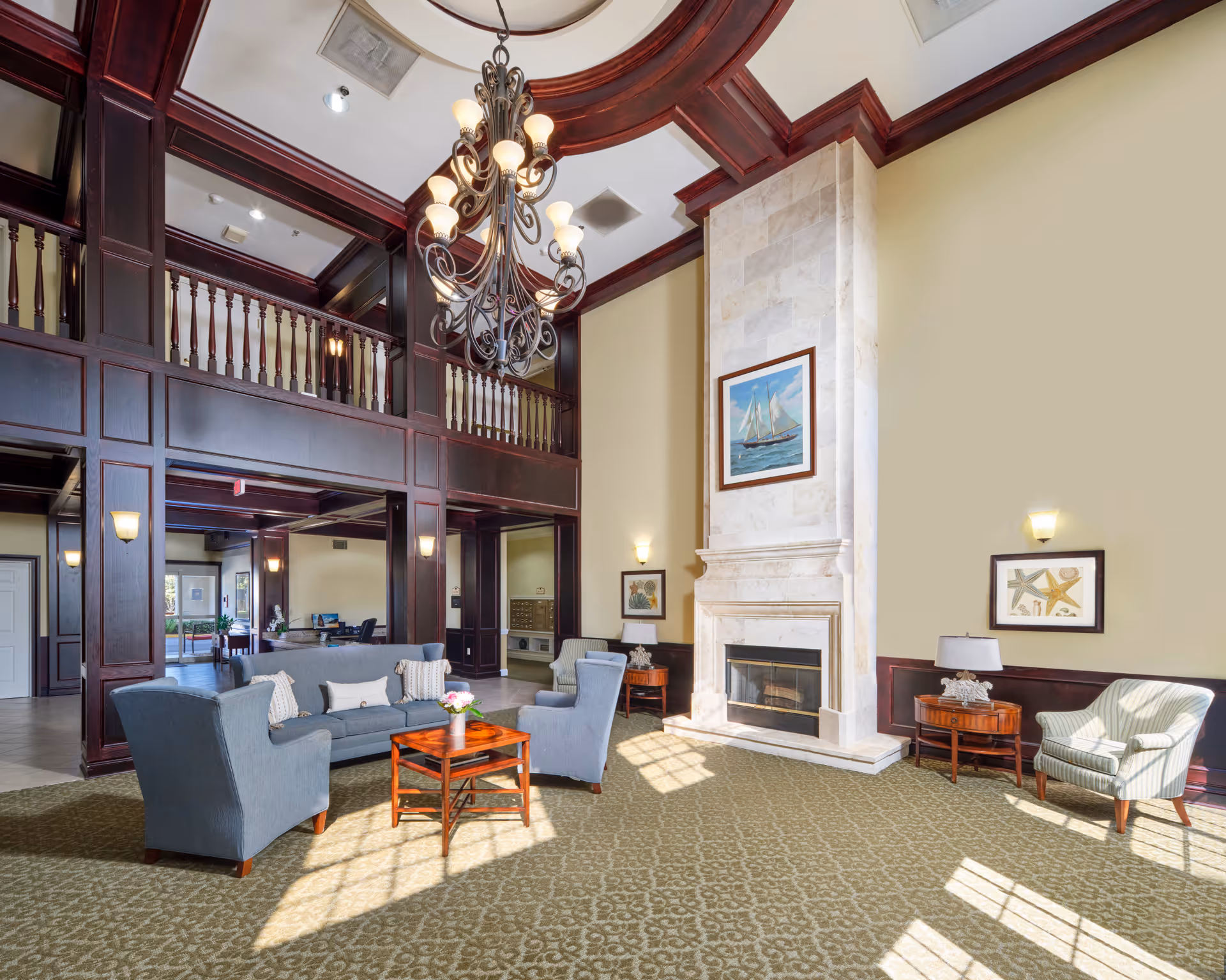 Bright, elegant common lounge with a high coffered ceiling and chandelier, a tall fireplace, and sofas and armchairs arranged on a patterned carpet beneath a wooden balcony.