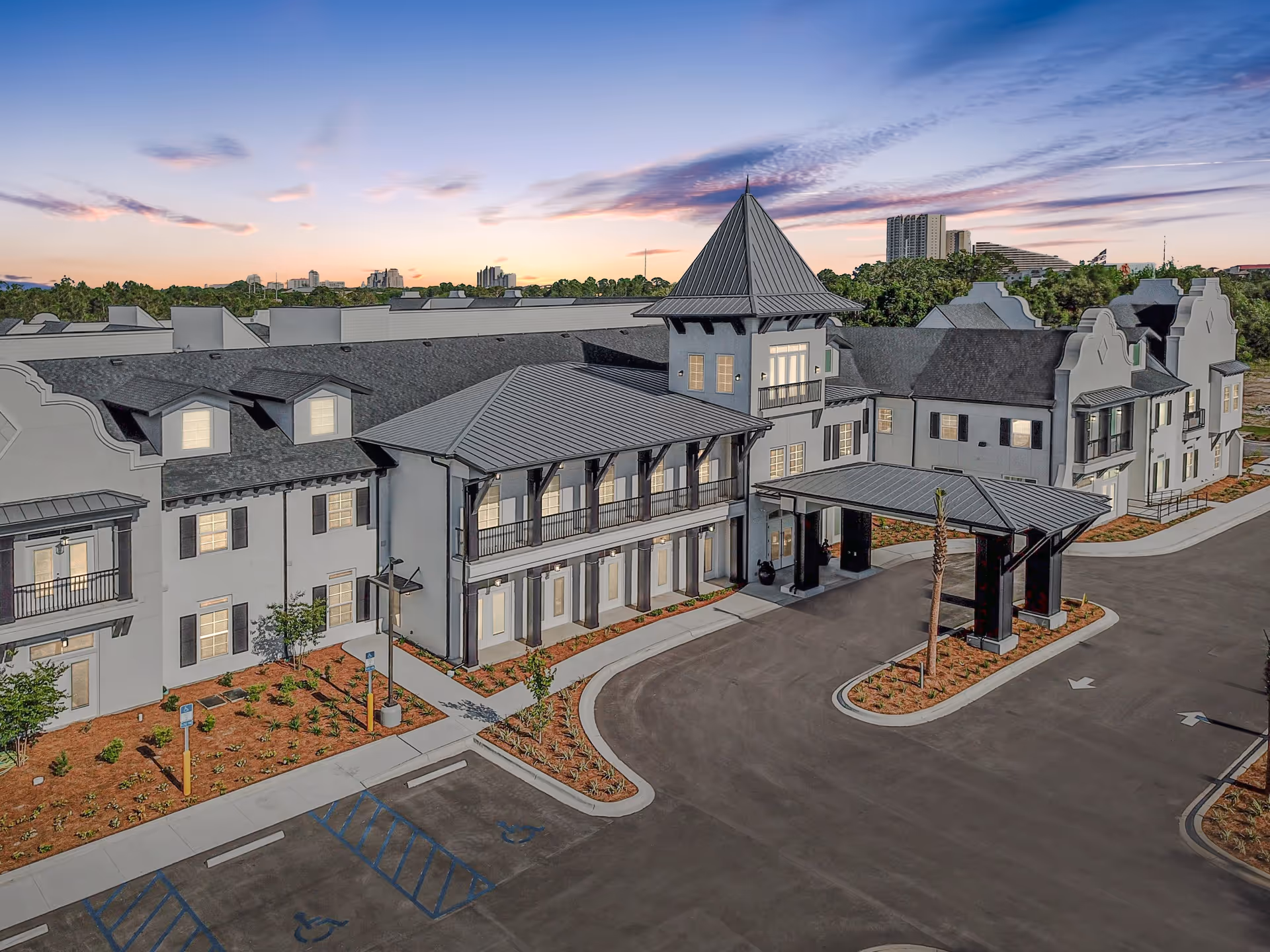 Exterior view of The Blake at Miramar Beach senior living facility at sunset, showing a large building with multiple windows, a covered entrance, landscaped areas, and a parking lot with handicap spaces.