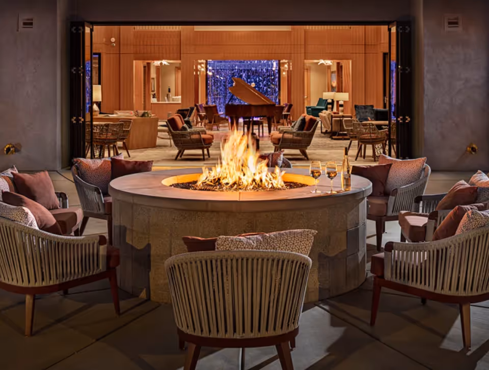 Circular outdoor fire pit surrounded by cushioned chairs on a patio opening into a warm indoor lounge with a grand piano.